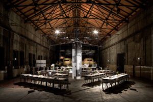 Eine alte Industriehalle in der provisorisch eine atmosphärische Beleuchtung installiert wurde und einige Tische in großem Quadrat angeordnet wurden. Im Hintergrund ist eine Küchenzeile erkennbar.