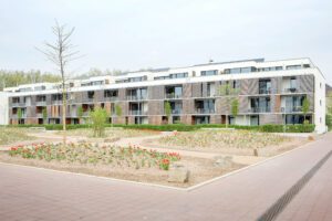 Ein modernes Gebäude mit drei Etagen und Balkonen. Im Vordergrund ist ein großes Blumenbeet und Bäume erkennbar.