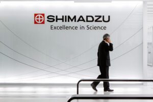 Eine Person in schwarzem Anzug telefoniert und läuft durch ein modernes Bürogebäude. Im Hintergrund ein Schriftzug "Shimadzu"