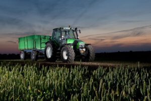 Ein Traktor mit Anhänger steht auf einem Acker, künstlich beleuchtet. Im Vordergrund ist Getreide zu erkennen. Im Hintergrund viel Himmel mit einem blau-orangen Farbverlauf.