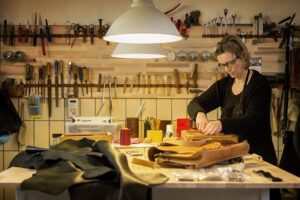 Eine Frau steht an einer Werkbank und arbeitet an Ledertaschen. Im Hintergrund sind zahlreiche Werkzeuge erkennbar.