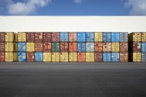 Zahlreiche Schiffs-Container stehen gestapelt vor einer weißen Lagerhalle vor blauem Himmel
